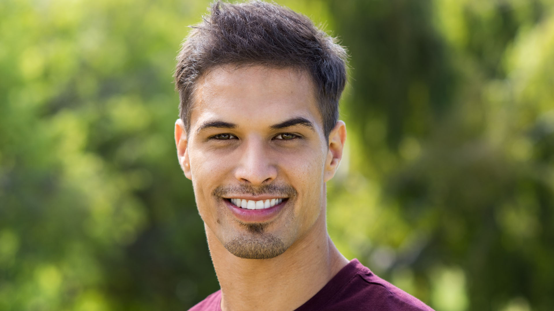 Young man with short dark hair and a light beard smiling outdoors, wearing a burgundy t-shirt with green trees behind him.